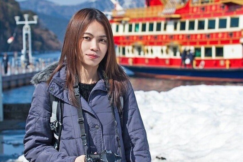 A person in a blue jacket with a camera strap, standing in front of a red and white boat on a body of water with snow on the ground. 3