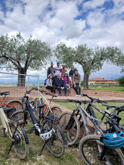 A group of people posing in front of bicycles parked on a grassy area with trees and a brick wall in the background. 3