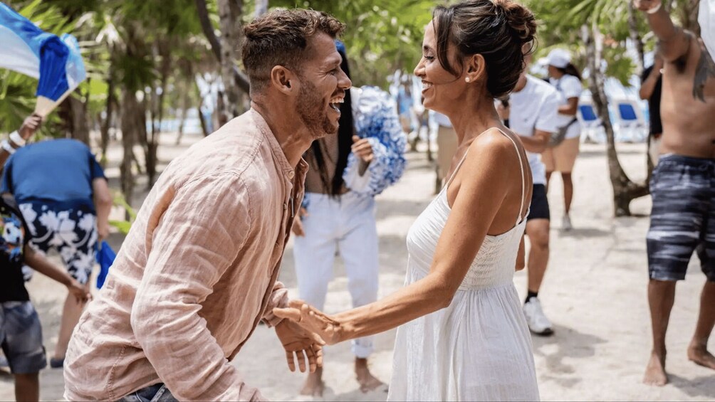 A couple dancing on a beach with palm trees and people in the background. 2