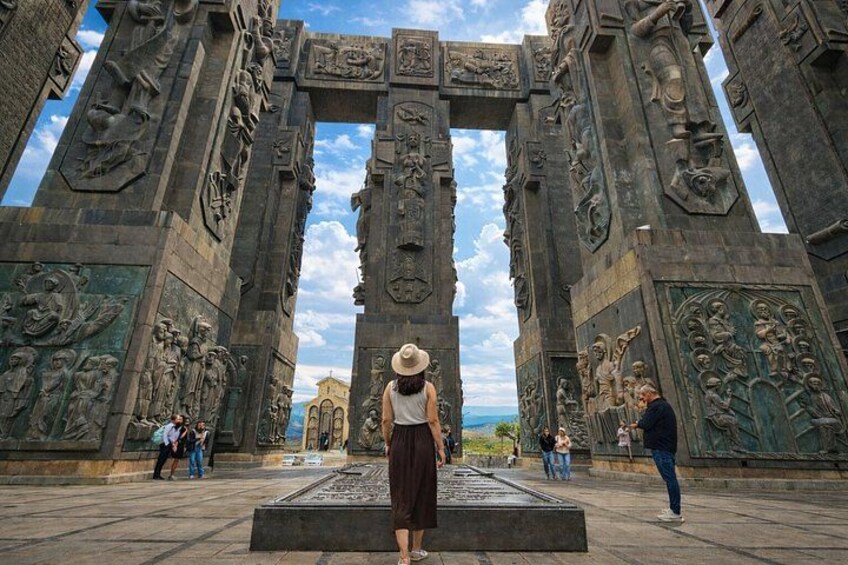 A large stone archway with intricate carvings, a person standing in the center, and a distant building. 1