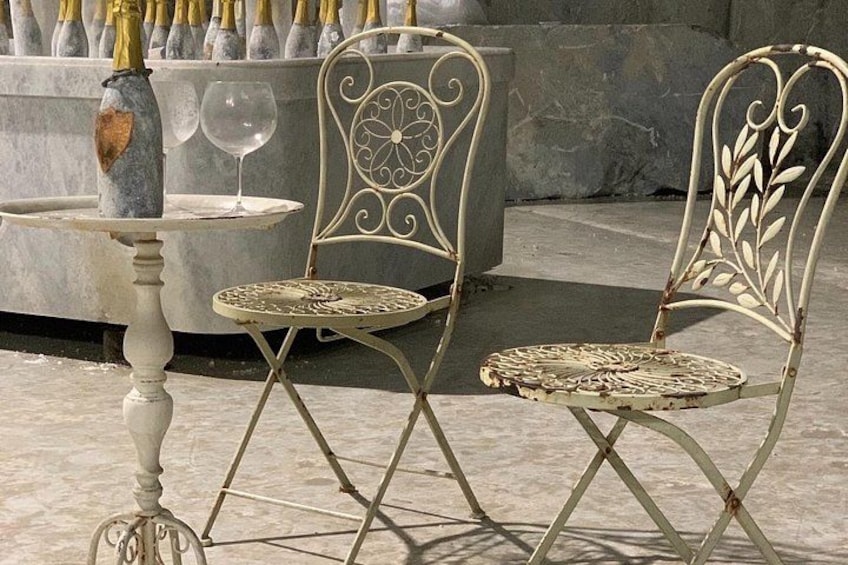 Two vintage metal chairs with intricate designs, a small round table, and a bottle of champagne with a glass. 1