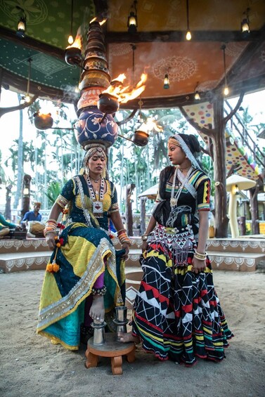 Two individuals in traditional attire, one balancing pots on her head, in an outdoor setting with hanging lights and a patterned wall. 4