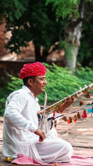 A person playing a traditional stringed instrument, seated outdoors with greenery in the background. 3