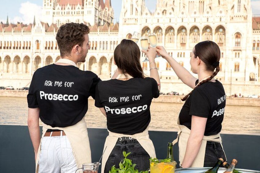 Tres personas con camisetas negras que tienen impreso en la parte de atrás el texto 'ASK ME FOR Prosecco', de pie junto a una masa de agua con un edificio grande y ornamentado al fondo. 1