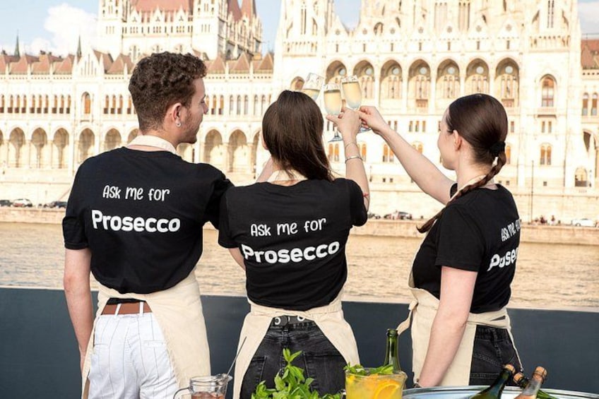 Drie mensen in zwarte t-shirts met de tekst 'ASK ME FOR Prosecco' op de achterkant, staan bij een rivier met een historisch gebouw op de achtergrond. 1