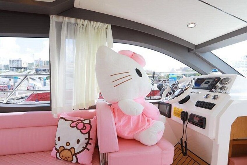 Interior de un barco con un peluche de Hello Kitty sobre un cojín rosa, un timón y vistas al puerto deportivo. 4