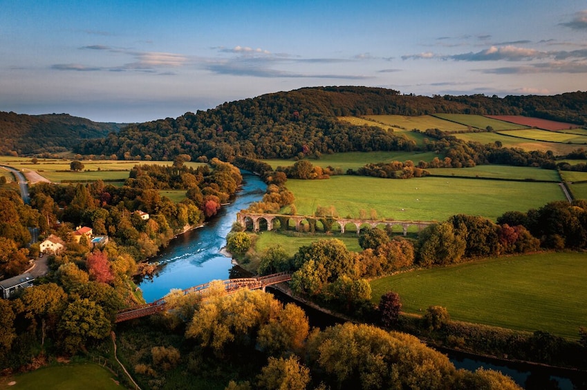 A river winding through a valley with fields, trees, and a stone bridge. 1