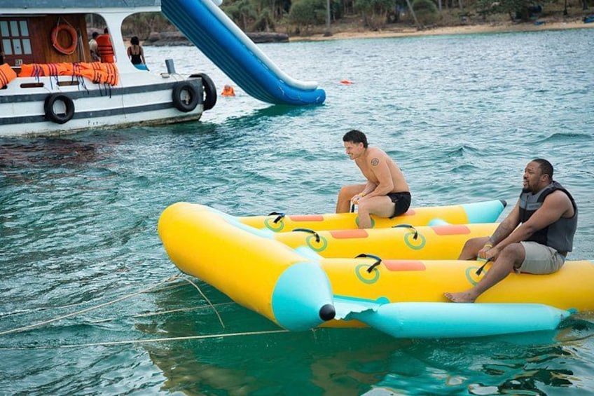Two people on a yellow inflatable raft in a body of water, with a boat and a blue slide in the background. 5