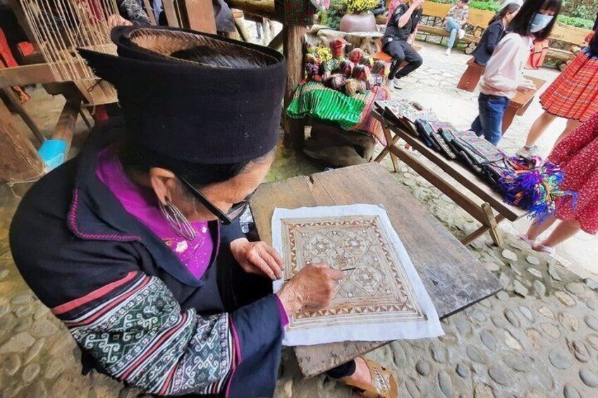 A person working on a patterned textile at a wooden table outdoors. 3