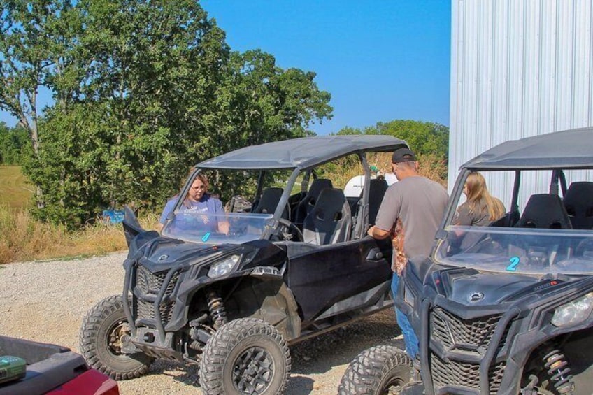 Two off-road vehicles parked on a gravel surface with a building and trees in the background. 4