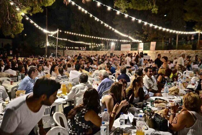 A crowded outdoor event with people seated at tables, string lights overhead, and a stone wall in the background. 5