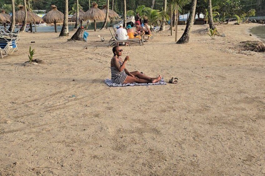 Una persona sentada sobre una toalla en la playa, con sombrillas y algunas personas al fondo. 3