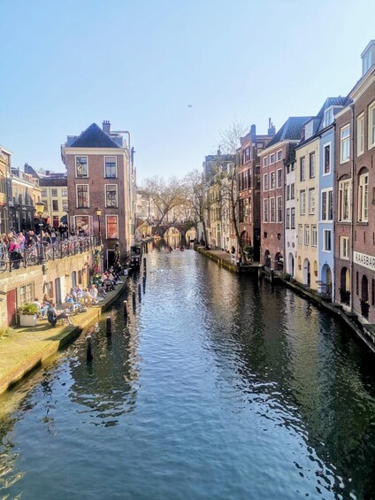 A canal flanked by historic buildings, a bridge in the distance, and people sitting by the water. 1