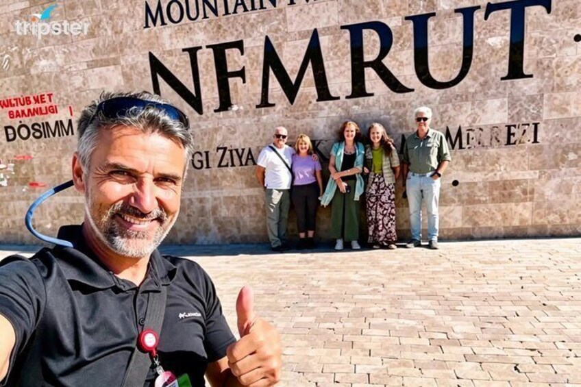 A man giving a thumbs-up in front of a wall with the text 'MOUNTAIN NFRUT' and a group of people standing behind him. 1