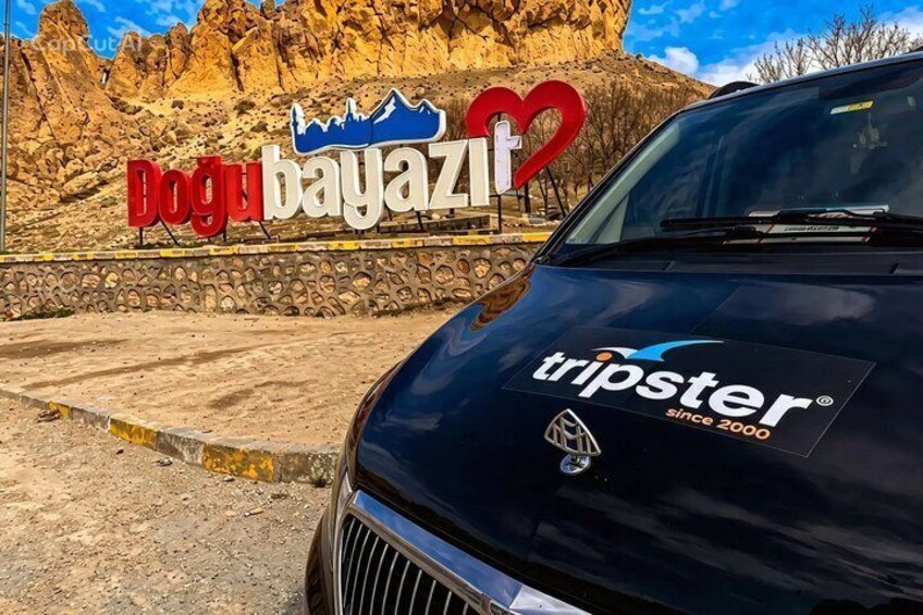A black vehicle with a Tripster logo parked in front of a sign that reads 'Dogu bayazit' with a mountain silhouette and a heart symbol. 4