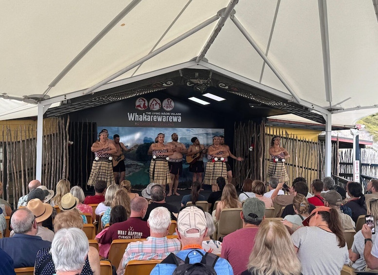 A stage with performers in traditional attire, an audience seated in front, and a backdrop with 'Whakarewarewa' text. 3