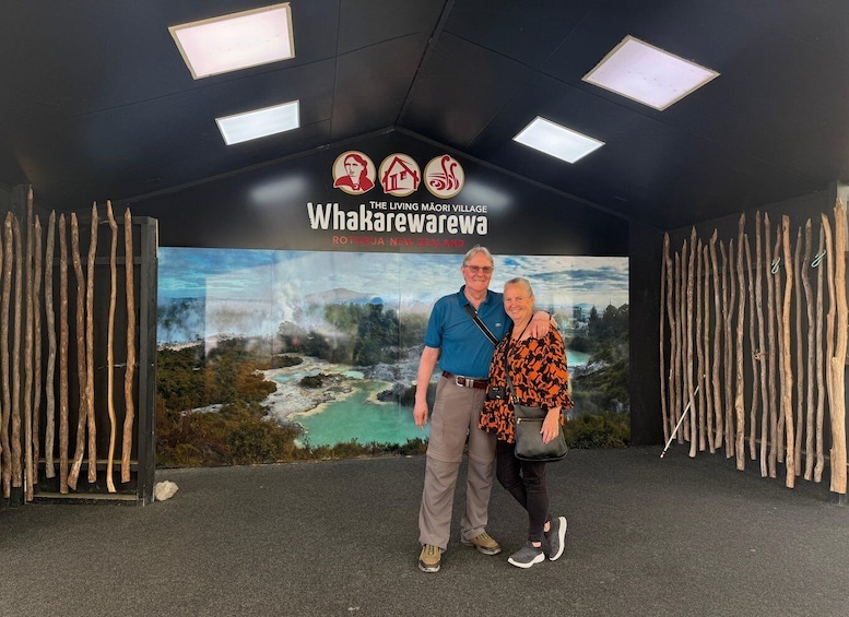 A man and woman standing in front of a sign for Whakaruru Village, surrounded by wooden stakes. 5