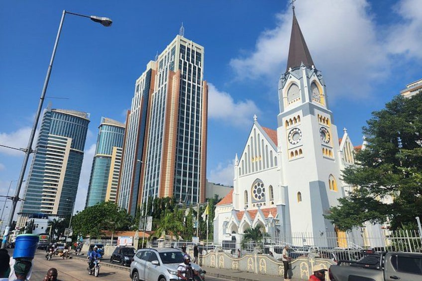 A church with a tall steeple and clock, surrounded by modern buildings and a busy street with cars and pedestrians. 4