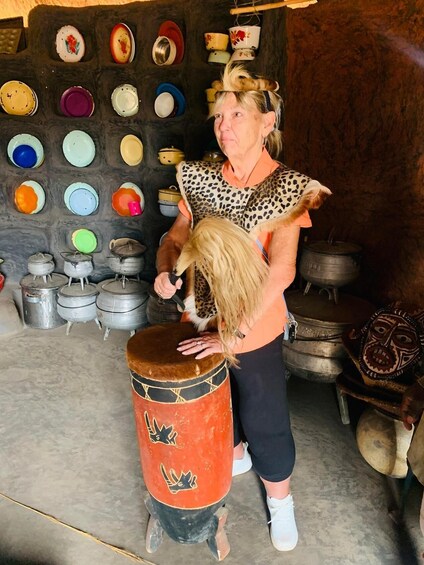 A person playing a drum in a rustic setting with various pots and dishes on the wall. 3