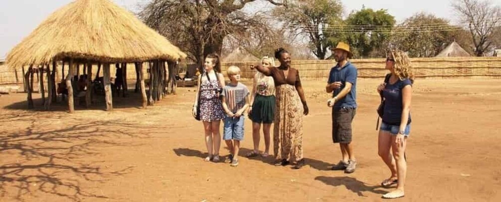 A group of people standing in a dry, open area with a thatched-roof structure in the background. 4