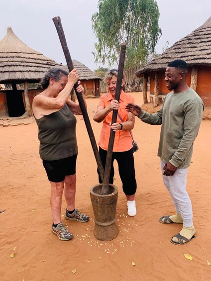 Three people interacting with a traditional wooden tool in a rural setting with thatched-roof huts. 1