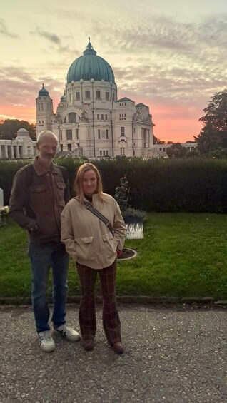 Two people standing in front of a large building with a dome, surrounded by greenery. 4