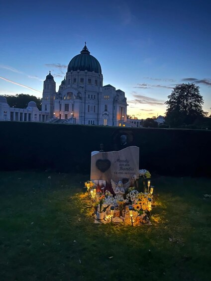A memorial with flowers and a plaque, set against a backdrop of a large, historic building at dusk. 1