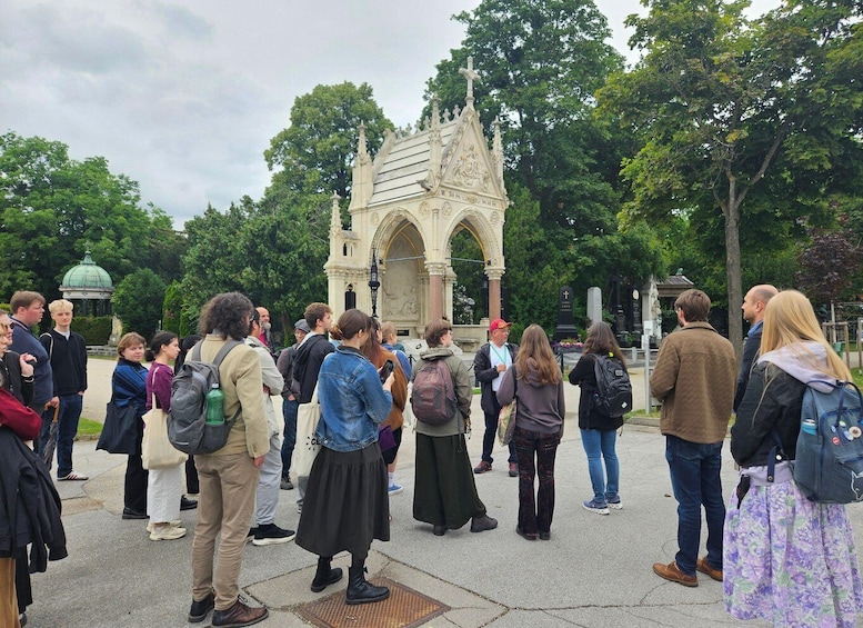 A group of people gathered around a Gothic-style monument in a park with trees and a dome in the background. 4