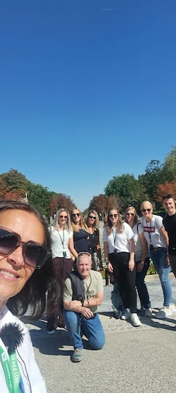 A group of people outdoors, some sitting and others standing, with trees and a clear sky in the background. 1