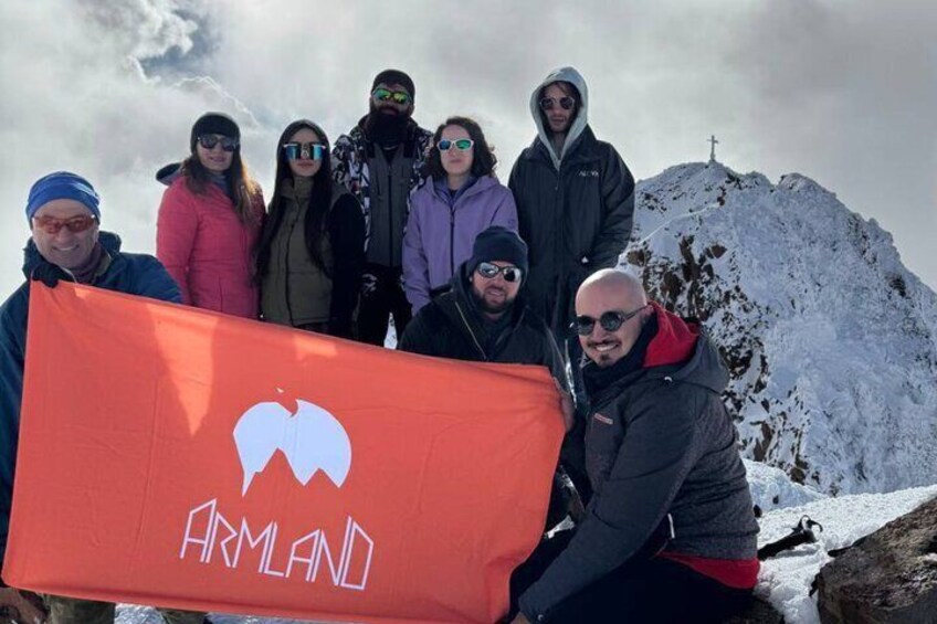 A group of people holding a flag with 'ARMLAND' written on it, standing on a snowy mountain with a cross on top. 1