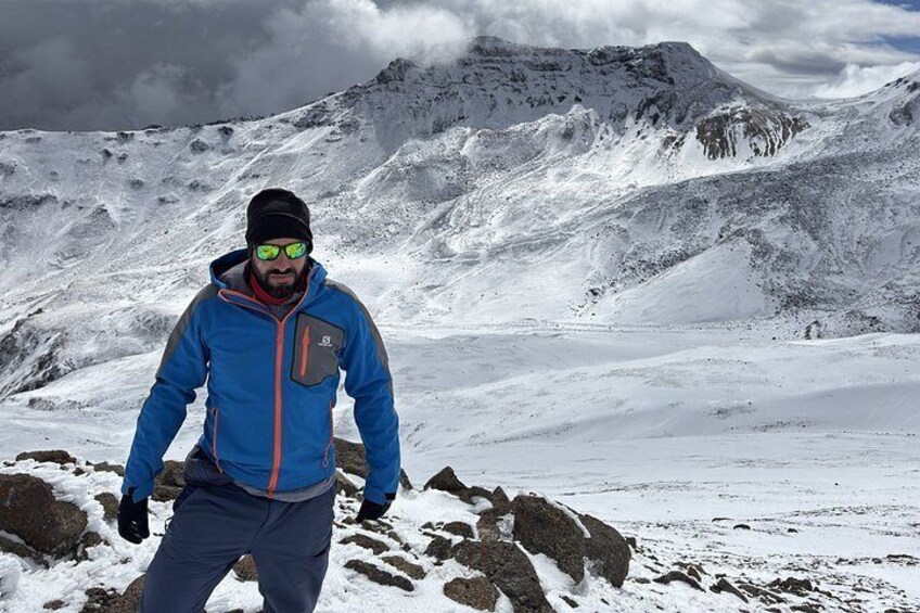 A person in a blue jacket standing on a snowy mountain with rocky terrain and a cloudy sky. 2