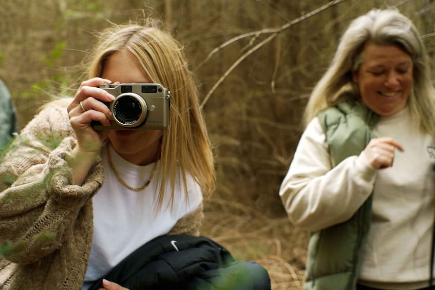 A person holding a camera, another person in a green vest, and a forested background. 2