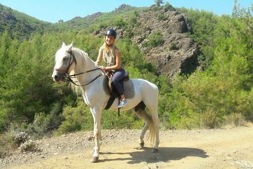 A person riding a white horse on a dirt path with green vegetation and rocky hills in the background. 3