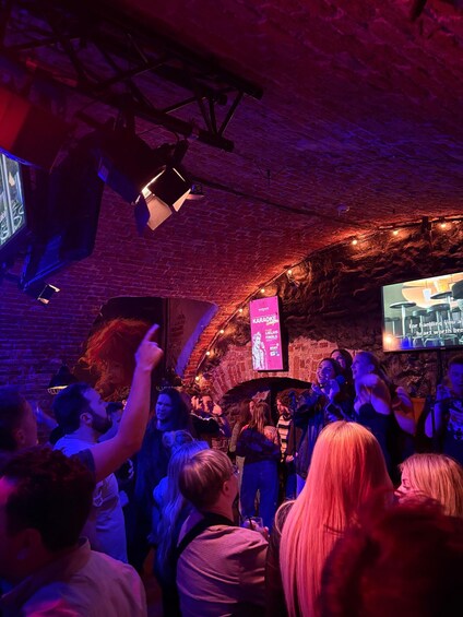 A crowd at a karaoke event in a brick-walled venue with a stage and a large screen displaying the event's name. 1