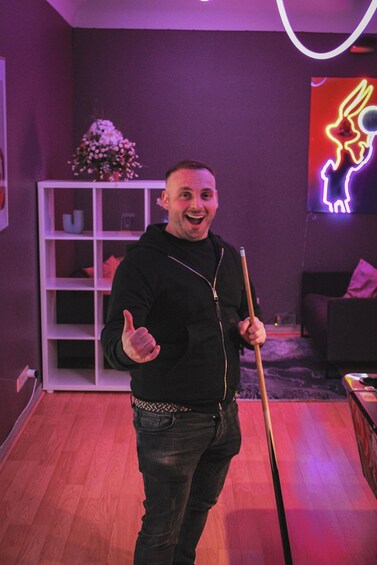 A person holding a pool cue in a room with purple lighting and a neon rabbit sign. 5