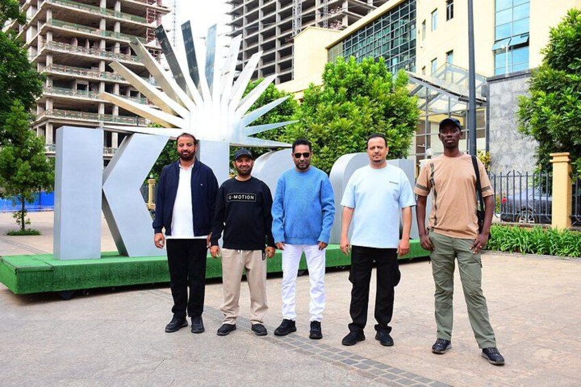 Four men standing in front of a large 'K' sign with a building in the background. 1