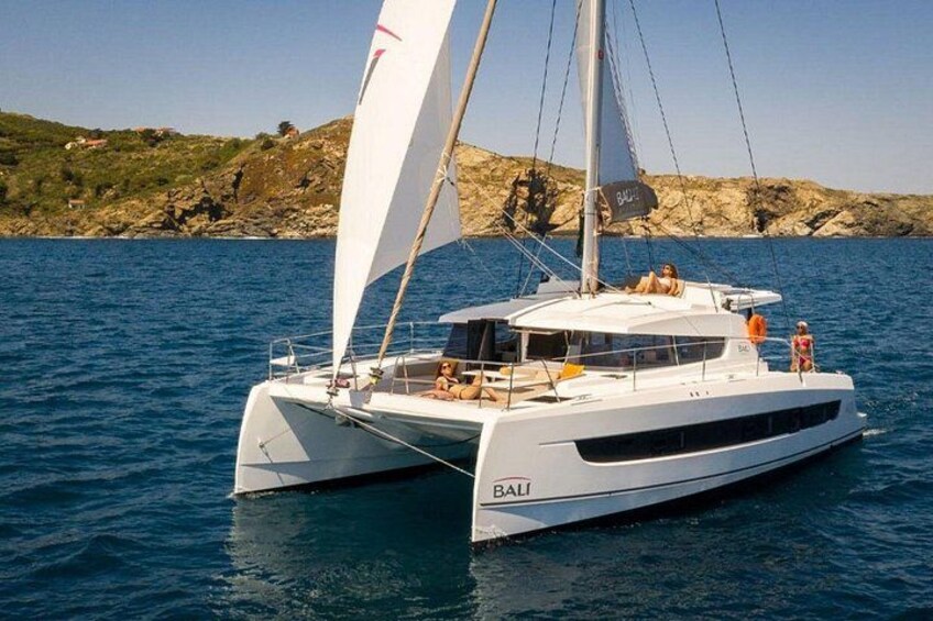 Un catamarán blanco llamado 'BALI' navegando sobre aguas tranquilas con una costa rocosa al fondo. 1