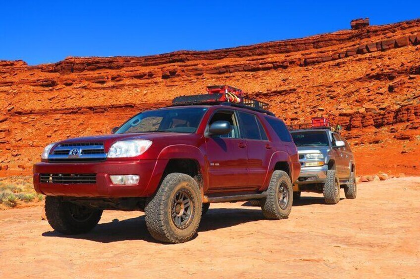 Dos vehículos SUV, uno rojo y otro plateado, estacionados en un camino de tierra con formaciones rocosas rojas al fondo. 4
