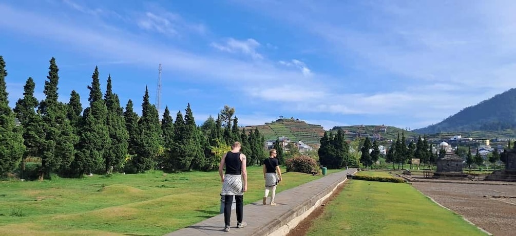 Two people walking on a path in a green landscape with trees and a hill in the background. 5