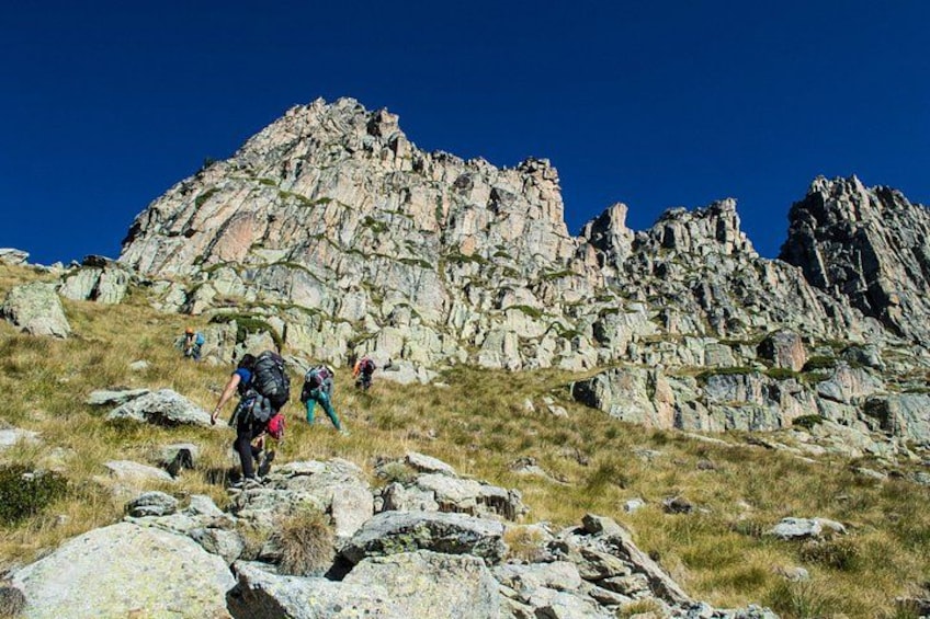 Hikers on a rocky mountain trail with large boulders and sparse vegetation. 3