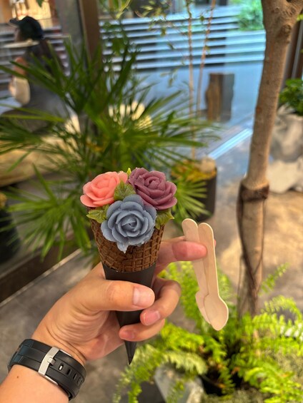 A hand holding a waffle cone with three ice cream roses in pink, purple, and blue. 1
