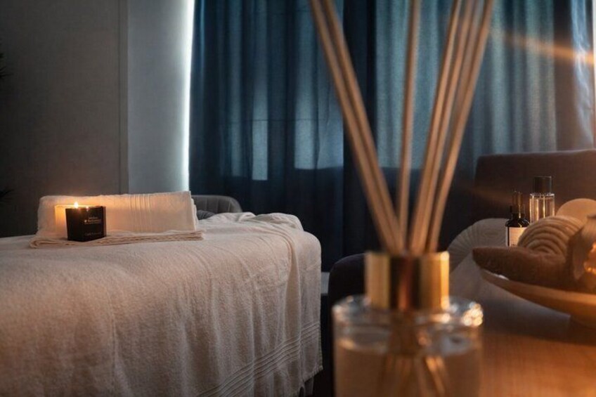 A hotel room with a bed, a lit candle, a vase with reed sticks, and a tray with towels. 1