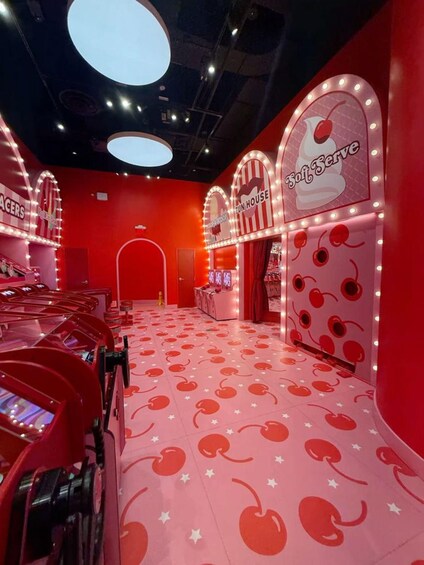 A brightly lit room with red walls and a floor decorated with cherry patterns. There are arcade machines and a sign that reads "Soft Serve." 1