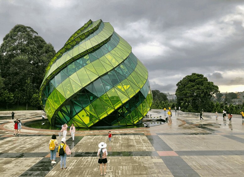 A large, green, spherical building with a reflective surface, surrounded by a plaza with people walking and a park in the background. 2