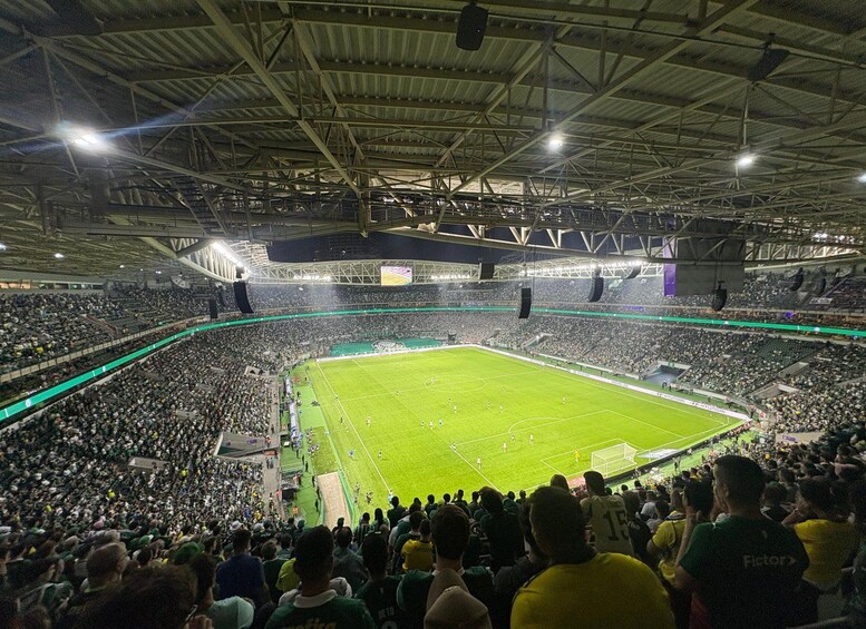 A large indoor stadium filled with spectators, a football field, and a high ceiling with metal beams. 1