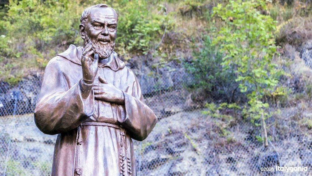 A statue of a man with a beard, wearing a robe, standing in a natural setting with trees and a fence in the background. 1