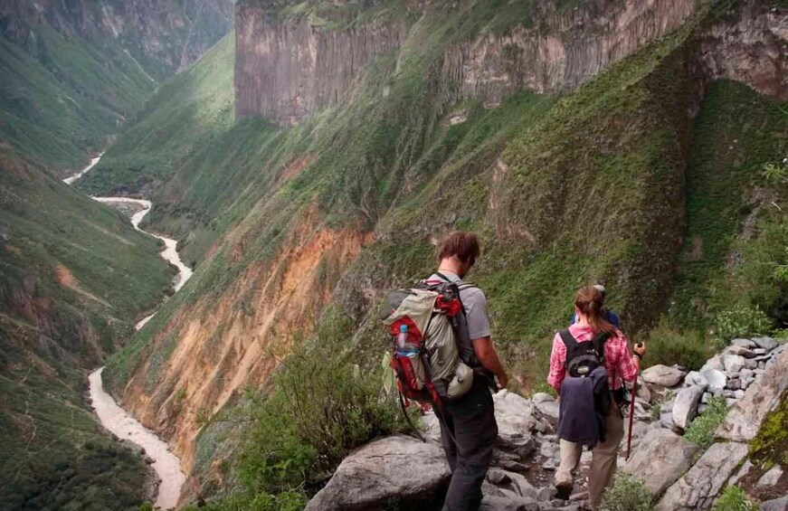 Two hikers on a rocky mountain trail with a river below. 1