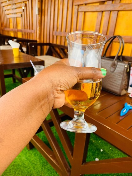 A hand holding a glass of beer with foam, a brown handbag, and a blue toy on a wooden table. 1
