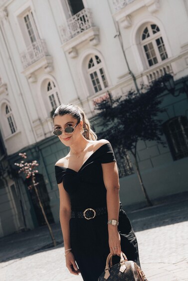 A person in a black dress with a belt, standing in front of a white building with arched windows and balconies. 4