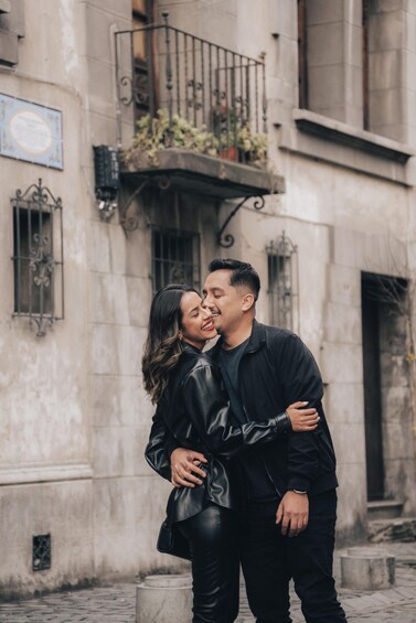 A couple embracing in front of a historic building with ornate ironwork balconies and a plaque. 1
